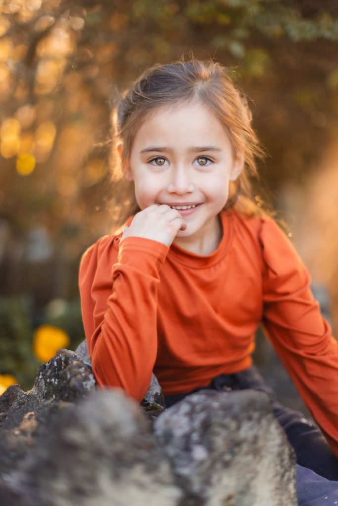 A child with brown hair wearing an orange longsleeve shirt sits outdoors, smiling with one hand near their chin. The background shows blurred green foliage, reminiscent of a charming portrait that could grace the walls of Belair Mansion.