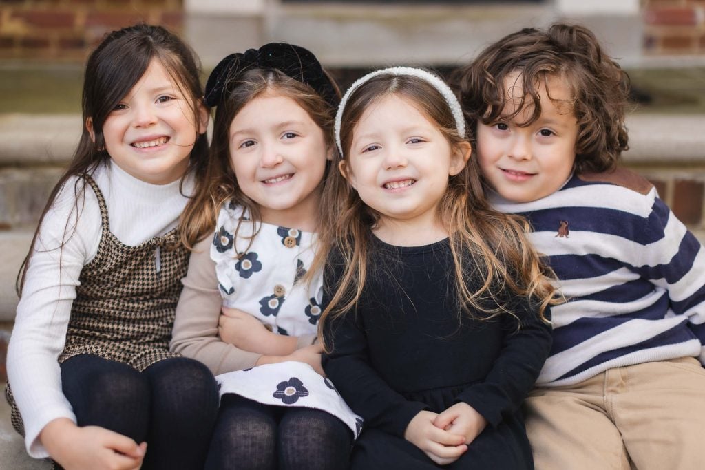 Four children are sitting together on the steps of Belair Mansion, smiling. The two girls on the left wear dresses with tights, the third girl wears a black dress, and the boy on the right wears a striped sweater and khaki pants. It's a timeless family portrait capturing pure joy.