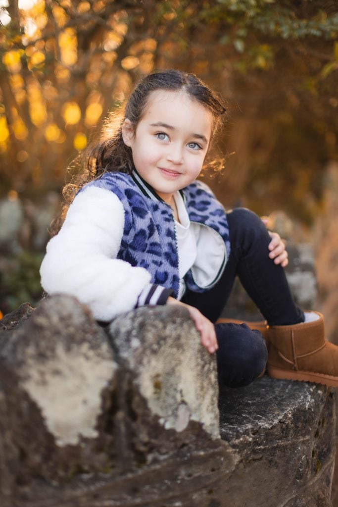 A child with long hair sits on a stone structure outdoors at Belair Mansion, wearing a white and blue jacket, black pants, and brown boots. The background features autumn foliage and warm light, creating the perfect setting for memorable portraits.