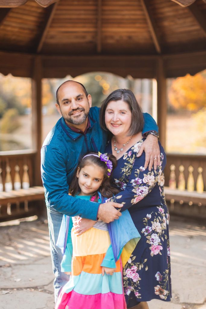 In a charming portrait at Greenspring Garden, a man and woman stand in a wooden gazebo, smiling and hugging a young girl in a colorful dress.