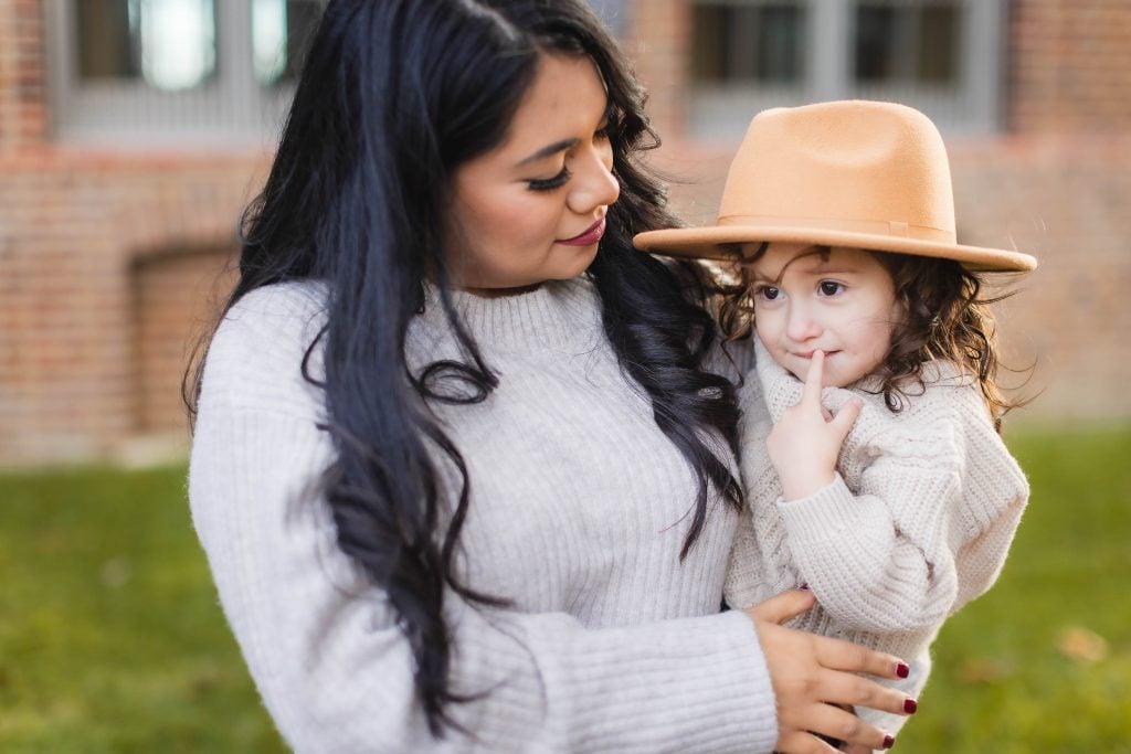 A family portrait shows a person holding a child wearing a tan hat and beige sweater. They are outdoors in front of the historic Belair Mansion.