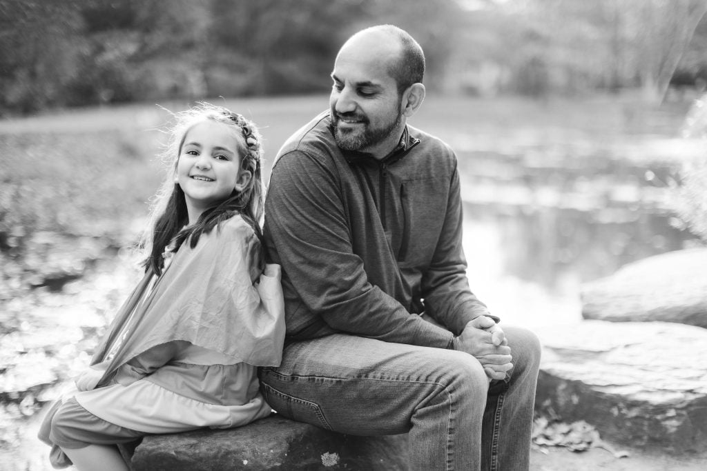 A bearded man sits on a rock near a body of water, looking at a smiling girl who is leaning against him. This portrait captures the warmth of family, as both enjoy the beauty of the outdoors in Alexandria's natural environment.