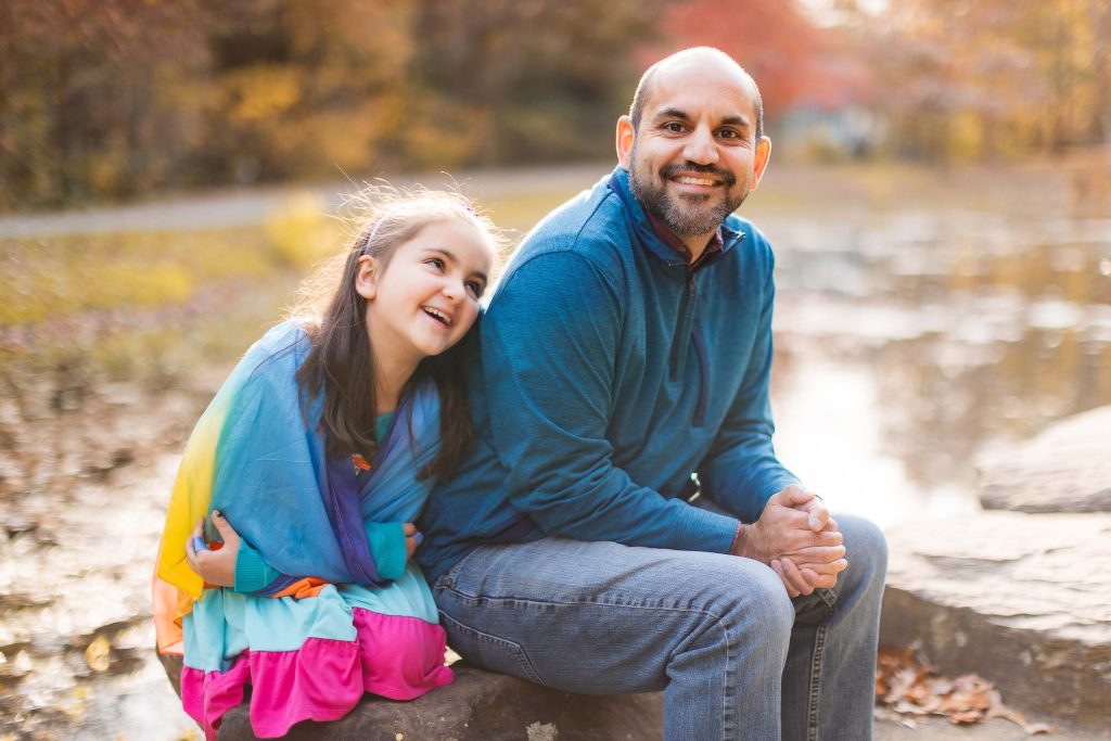 A man in a blue shirt and jeans sits on a rock near a body of water, with a young girl in a colorful dress resting her head on his shoulder. Both are smiling, creating a perfect family portrait. Trees and autumn foliage at Greenspring Garden form the picturesque background.