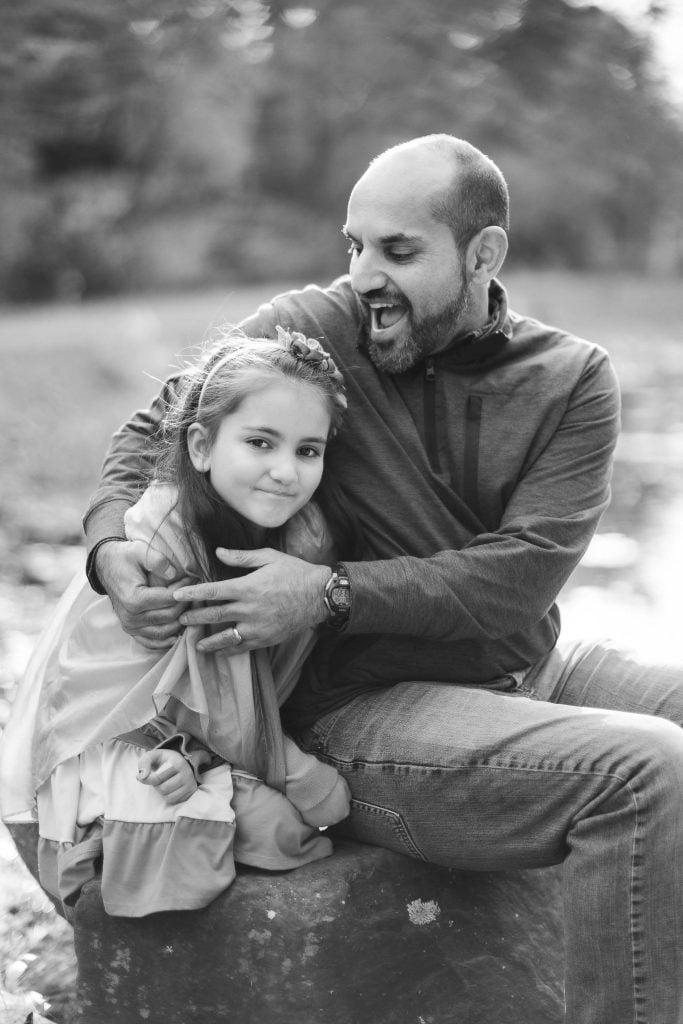 Black and white photo of a bearded man sitting on a rock, embracing a young girl. The man appears to be pointing to the distance while the girl smiles slightly. Trees and blurred background in the quaint town of Alexandria, Virginia, visible. The scene exudes warmth and family bonds.