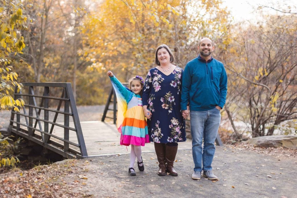 A family of three stands in front of a wooden bridge at Greenspring Garden in Alexandria, surrounded by autumn foliage. The young girl wears a colorful dress, while the woman and man sport casual fall outfits.