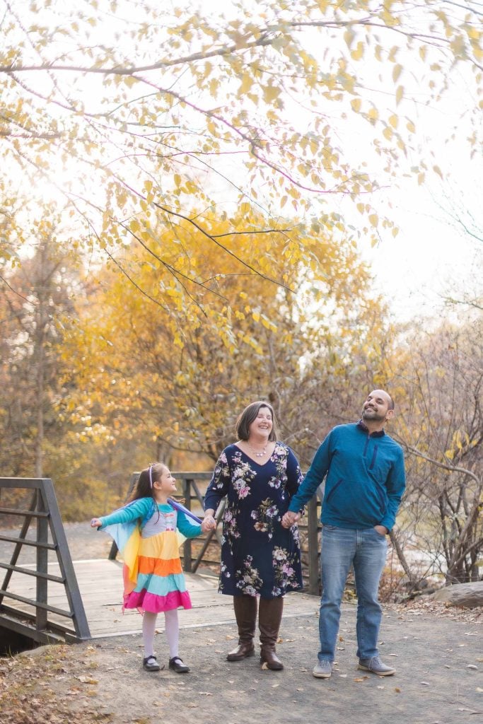 A family of three, including a child in a colorful dress, stand on a bridge at Greenspring Garden in Virginia surrounded by autumn trees. They are holding hands and looking up at the foliage.