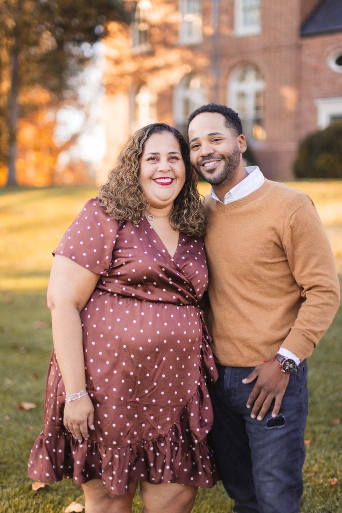 A woman in a brown polkadot dress and a man in a tan sweater stand smiling together outdoors with the historic Belair Mansion and fall foliage in the background.