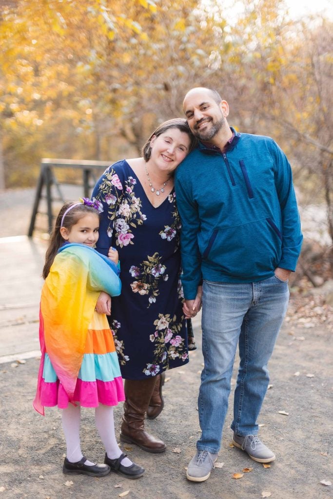 At Greenspring Garden, a woman, a man, and a girl stand together on a path. The woman and girl wear colorful dresses while the man sports a blue pullover. The girl proudly dons her rainbow cape as they all smile for the portrait amidst the blooms of Alexandria.