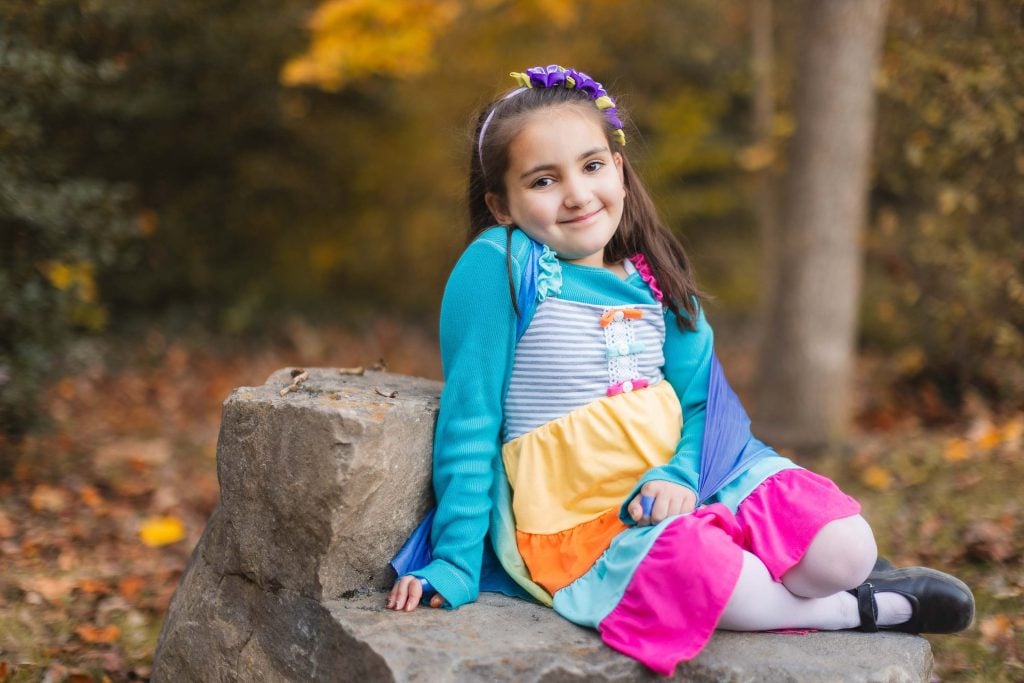 A young girl named Alexandria, wearing a colorful dress and a flower headband, sits on a rock surrounded by the vibrant fall landscape of Virginia.