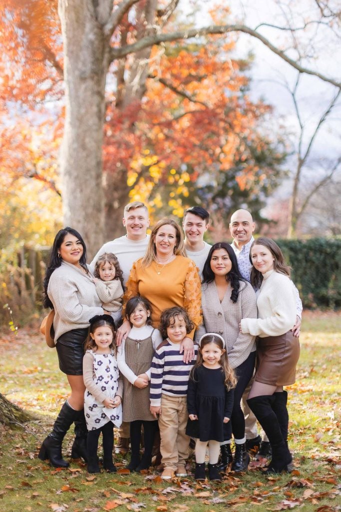 Group photo of eleven people in a park during fall, composed of adults and children. They are smiling and standing closely together, with autumn leaves visible on the ground and trees in the background. It captures a warm family portrait that feels straight out of Belair Mansion's picturesque setting.