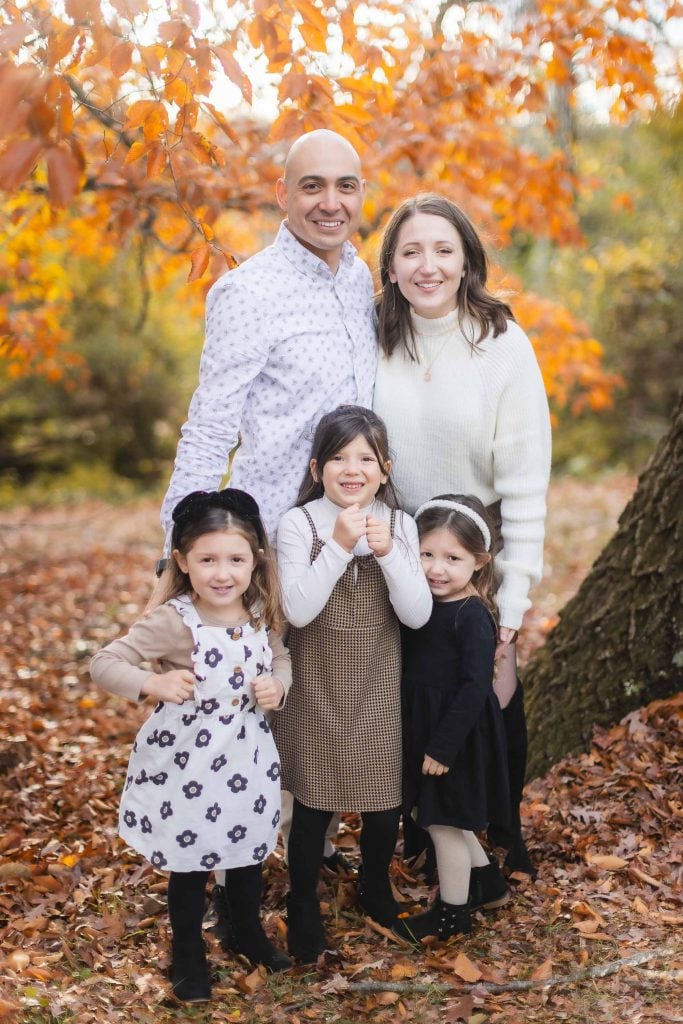 A family of five stands outdoors in an autumn setting, with three children and two adults smiling at the camera amidst fallen leaves and colorful foliage, creating a perfect portrait against the backdrop of Belair Mansion.