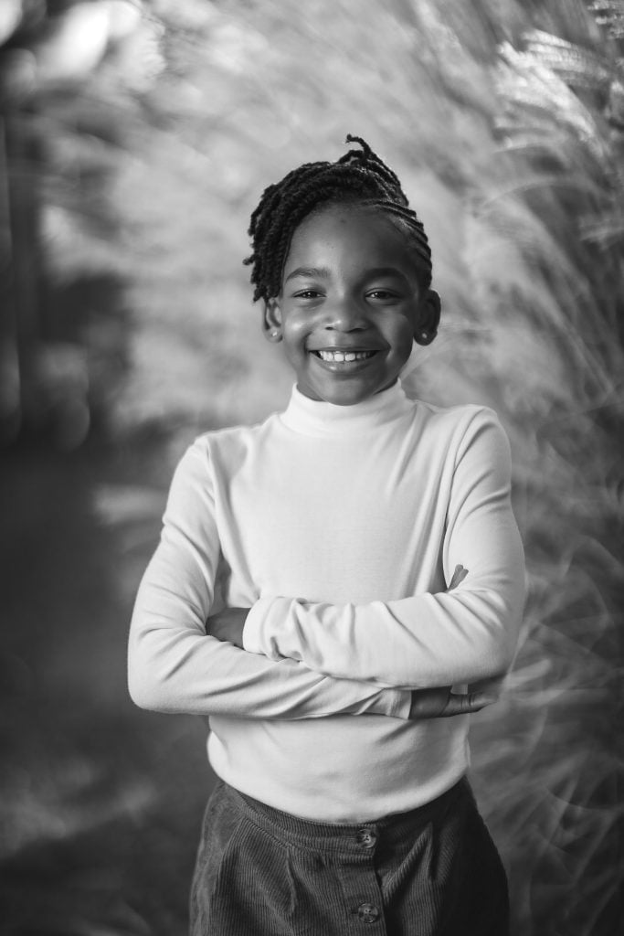 Black and white family portrait of a young person with a braided hairstyle, wearing a longsleeve shirt, standing with arms crossed and smiling at the camera.