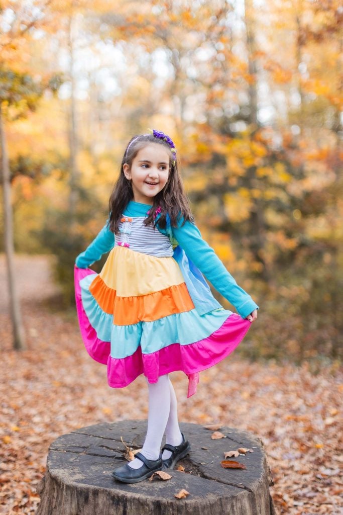 In this portrait, a young girl stands on a tree stump in a colorful, layered dress, holding the sides of her outfit. The background showcases the vibrant autumn foliage of Greenspring Garden's forest.