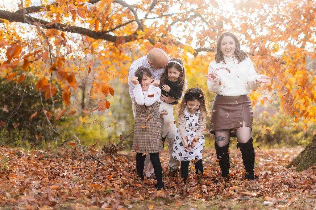 A family of five stands outdoors among autumn leaves. The mother and father, along with their three daughters, are enjoying the fall weather at Belair Mansion. The children are playing with the fallen leaves, creating a memorable family portrait in the picturesque setting.