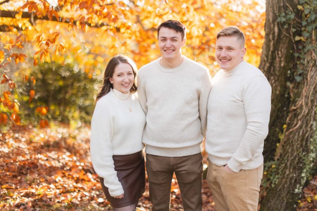 Three individuals standing outdoors in autumn, wearing lightcolored sweaters and smiling, with colorful fall foliage in the background at Belair Mansion—it's a heartwarming family portrait.