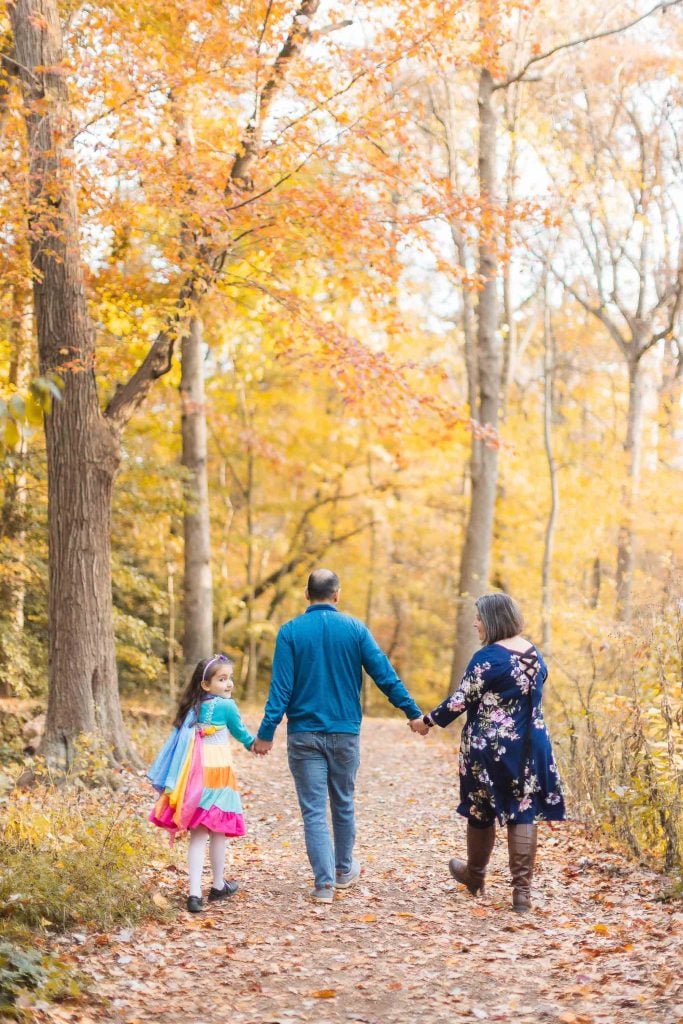 A family of three holding hands, walking down a forest path lined with autumncolored trees in Greenspring Garden, Alexandria. The child is dressed in a vibrant, colorful outfit.