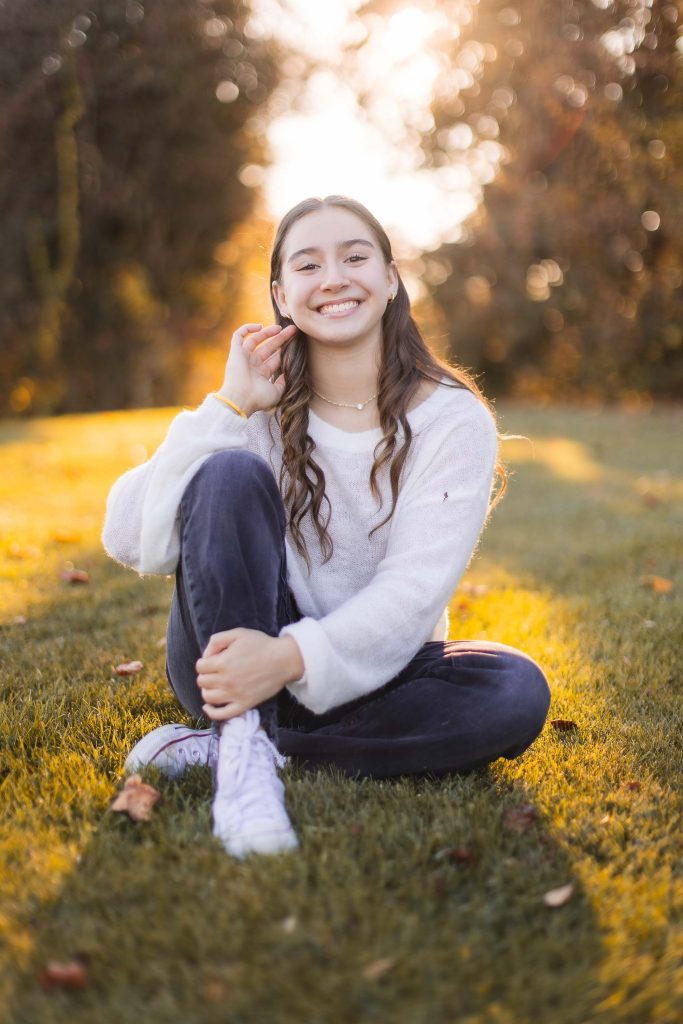 A person with long hair sits on grass in a park near Belair Mansion, smiling. They wear a white sweater, dark jeans, and white sneakers. The background features trees and sunlight shining through.