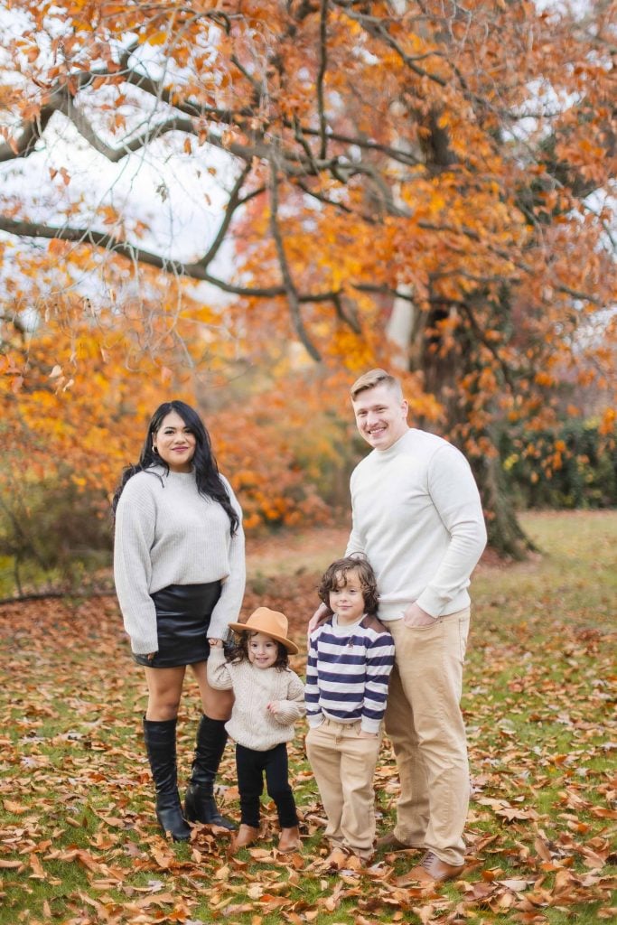 A family of four stands outdoors in autumn at Belair Mansion. The woman and man pose with two children; one child is wearing a hat. They are surrounded by fallen leaves and trees with orange foliage, creating a perfect family portrait.