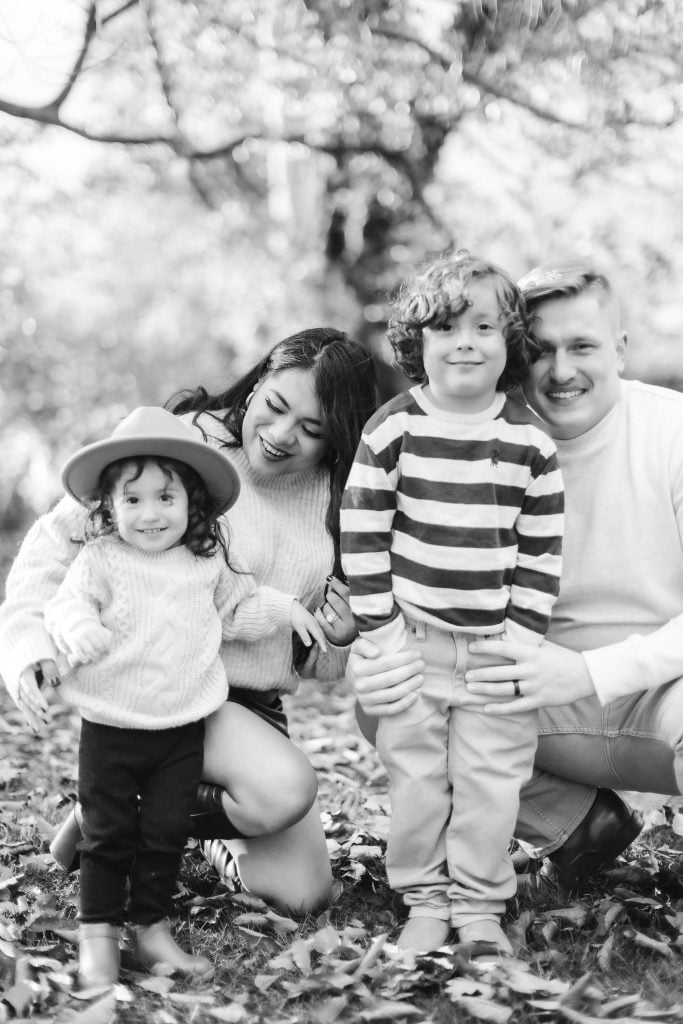 A black and white family portrait with two young children outdoors. The parents kneel while the children stand in front, surrounded by trees and fallen leaves, reminiscent of a Belair Mansion scene. The girl wears a hat and the boy has curly hair.