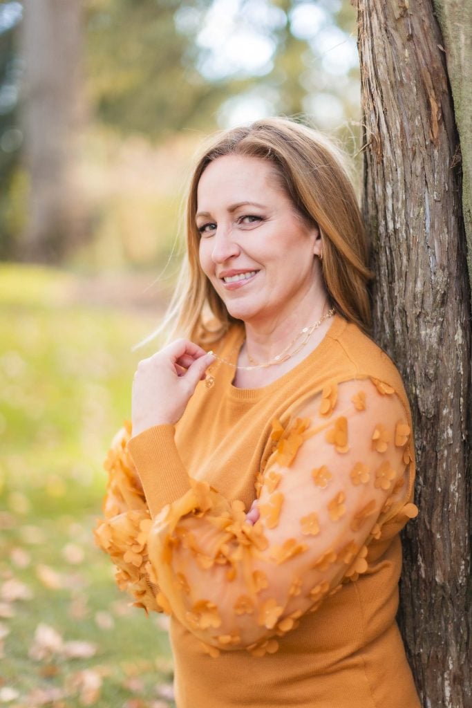 A woman in an orange sweater with floral sleeves leans against a tree outdoors, smiling, as if posing for a family portrait. The background features blurred greenery, reminiscent of the serene gardens at Belair Mansion.