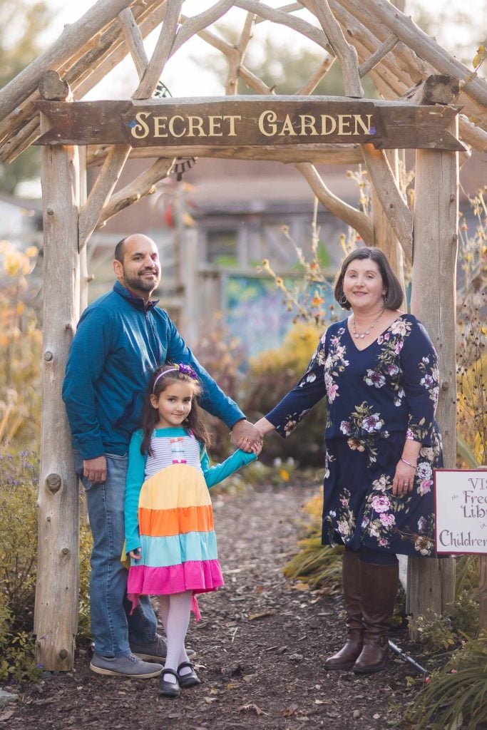 A portrait of a man, young girl, and woman stands beneath a wooden archway labeled "Secret Garden" in the serene setting of Greenspring Garden, Virginia. They hold hands, their smiles reflecting the beauty and tranquility surrounding them.