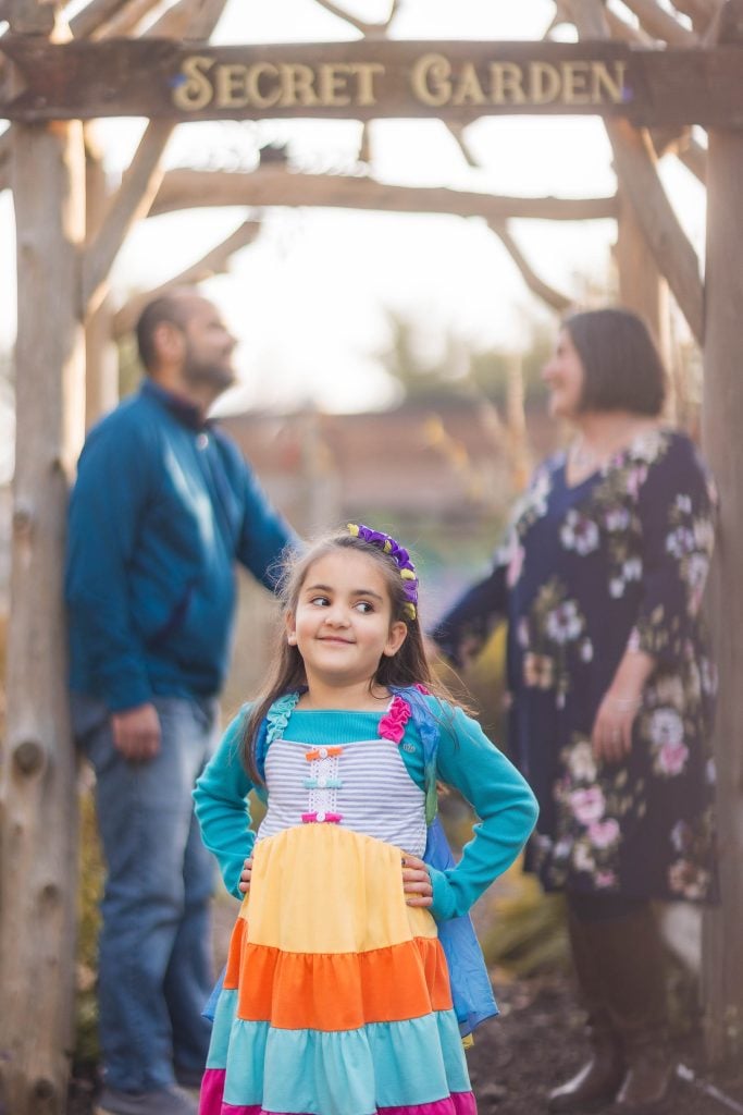 A young girl in a colorful dress stands in the foreground. Behind her, a man and woman stand under a wooden archway with a "SECRET GARDEN" sign, capturing the charm of Greenspring Garden in Alexandria.