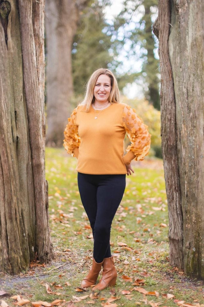 A portrait of a woman in an orange sweater and black pants stands between two large tree trunks on a grassy area with fallen leaves at Belair Mansion.