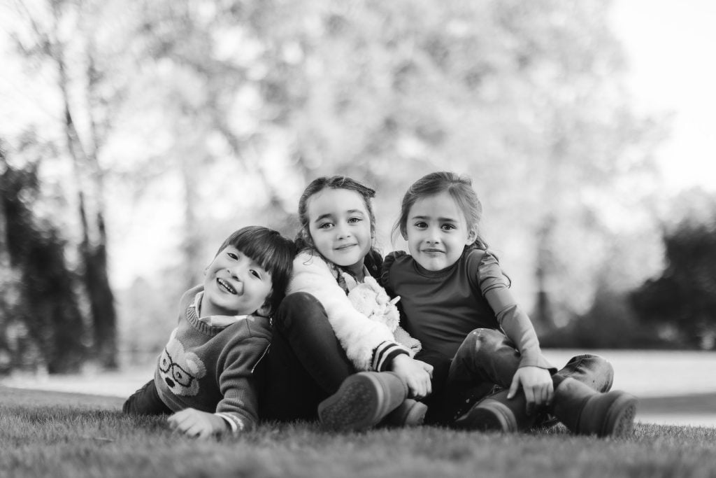 Three children sit on the grass in an outdoor setting, smiling and looking at the camera. The background is a blurred natural landscape that adds to the charm of this family portrait taken near Belair Mansion.