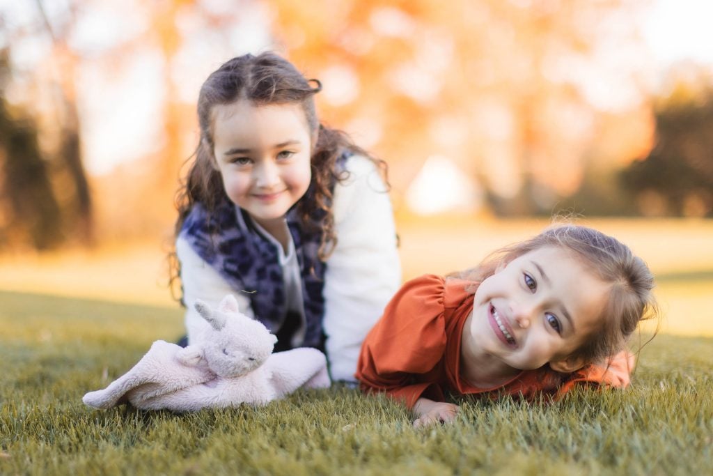 Two young girls lie on the grass with a plush toy, smiling at the camera, creating a heartwarming scene. The background is filled with blurry, autumncolored trees near Belair Mansion, making it a perfect setting for family portraits and extended family gatherings.