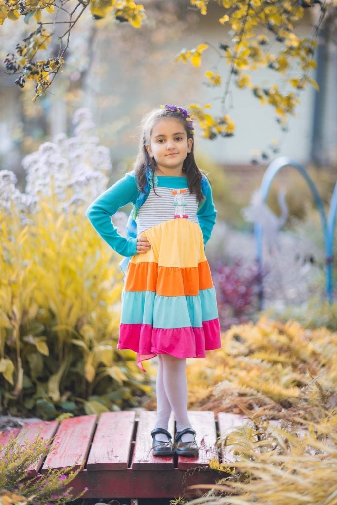 A young girl stands on a red wooden pathway in Greenspring Garden, Virginia, wearing a colorful striped dress and teal cardigan, with her hands on her hips and an autumn background.