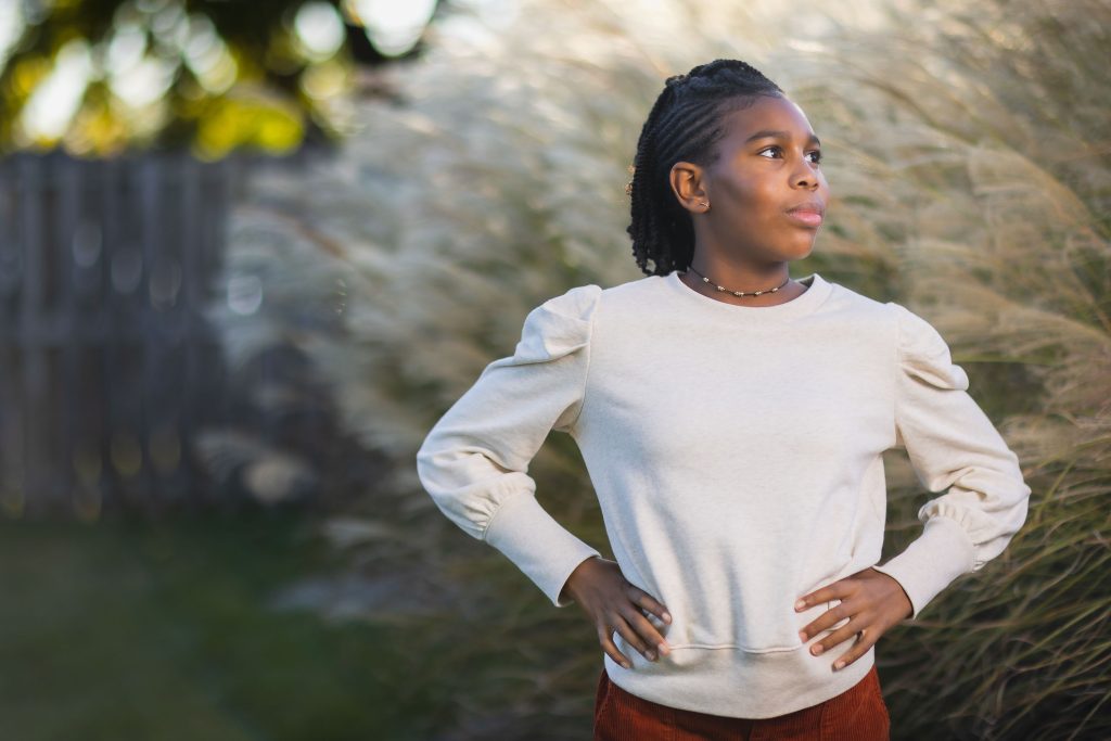 A person stands outside with hands on hips, wearing a lightcolored sweatshirt and looking to the side. Tall grass and a wooden fence are in the background, creating a serene setting perfect for a family portrait.