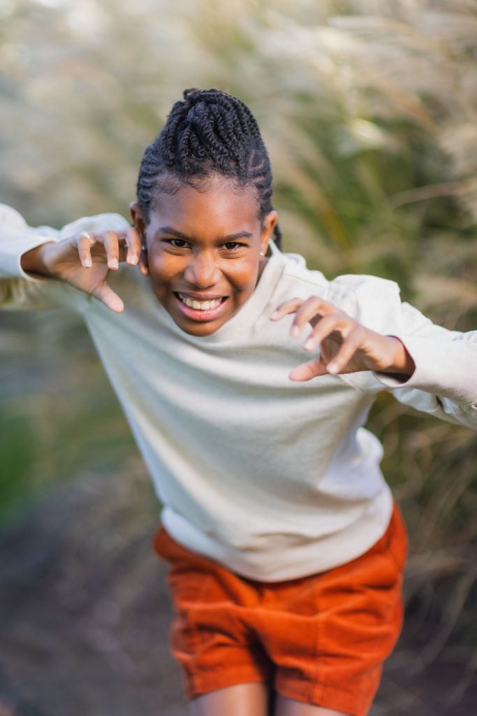 A portrait of a child with braided hair, wearing a white sweater and orange shorts, playfully poses with raised hands outdoors—a perfect snapshot for the family album.