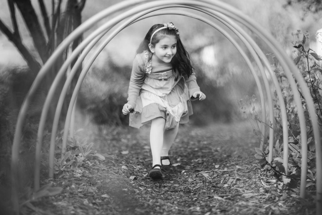 A young girl in a dress and headband crawls through a circular garden arch structure on a dirt path in Alexandria, smiling. The photo is in black and white.