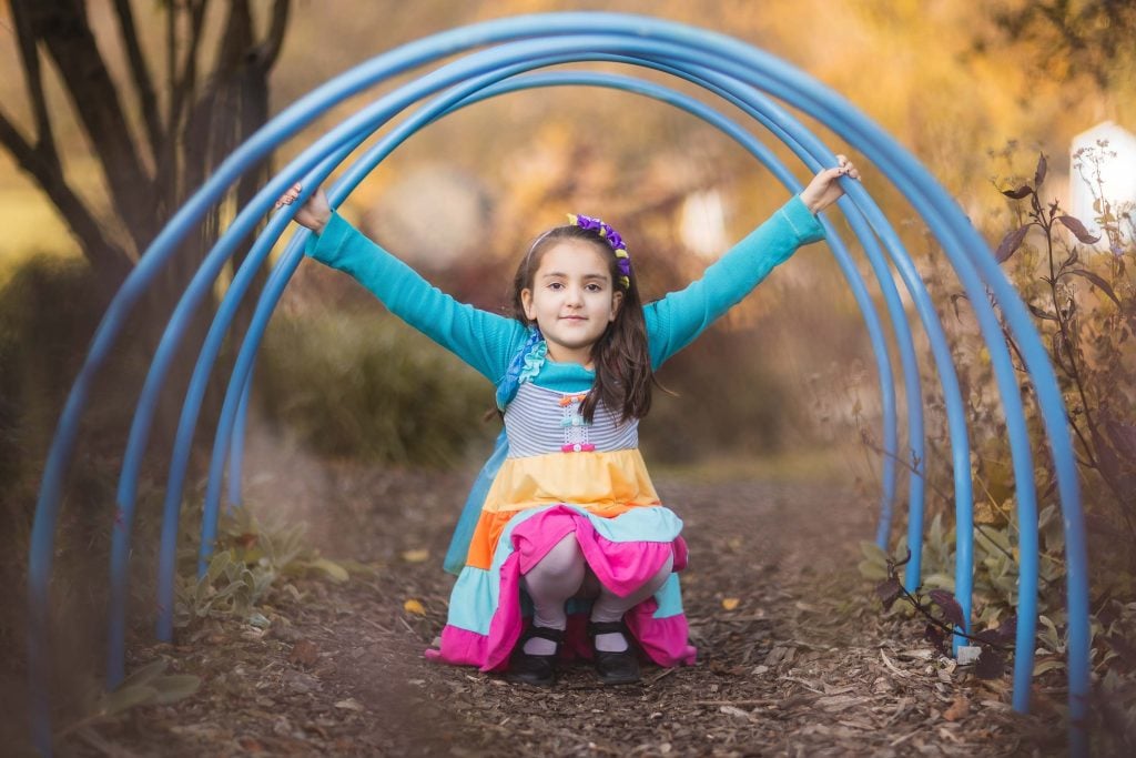 A young girl with a colorful dress and blue sweater squats under blue metal arches at Greenspring Garden in Alexandria, holding onto the arches with both hands.