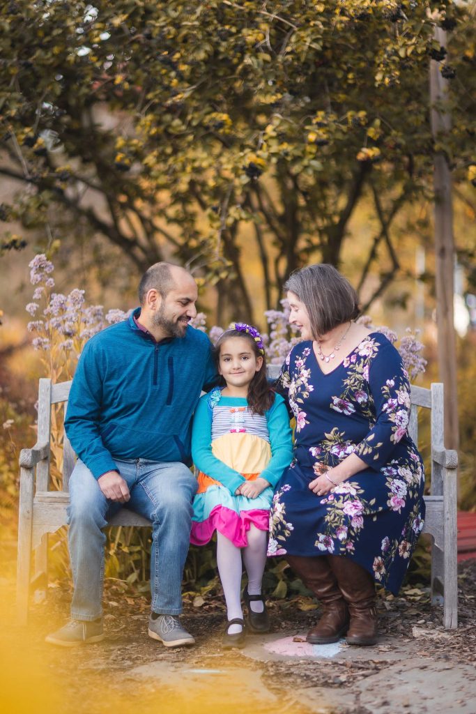 In the garden of their Alexandria home, a family of three sits on a bench. The child in colorful clothing is in the middle, with the man on the left and the woman on the right. All are smiling and dressed casually, creating a perfect portrait of happiness in Virginia.