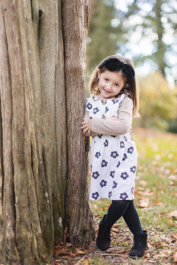 A young girl in a white dress with black flowers leans against a tree. She wears black leggings, boots, and sunglasses on her head. The background features a park with autumn foliage, evoking the charm of a family portrait set near the stately Belair Mansion.