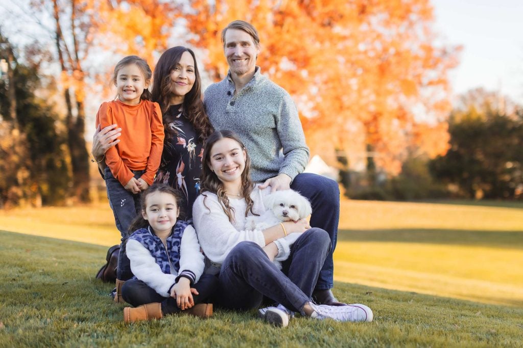 A family of five, including two adults and three children, pose outdoors on a lawn with fall foliage in the background at Belair Mansion. One child holds a small white dog.