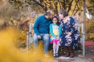 A man, a woman, and a young girl sit closely on a wooden bench at Greenspring Garden, surrounded by autumn foliage. The adults lean in affectionately towards the girl, who smiles warmly in her colorful dress. This family portrait captures the essence of loving connections and nature's beauty.