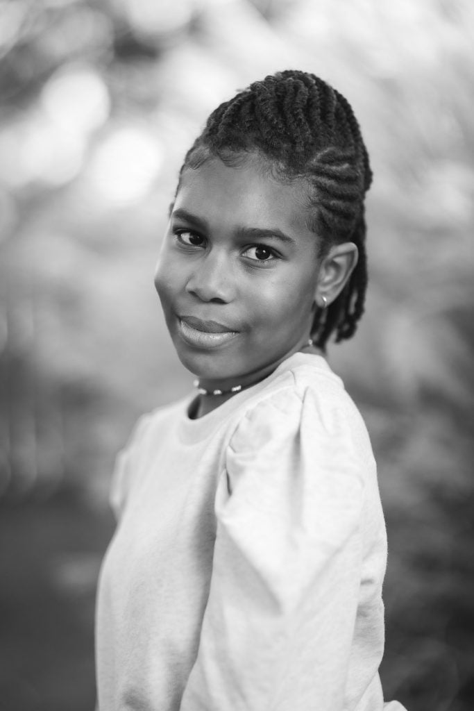 Black and white portrait of a young girl with braided hair, wearing a longsleeved shirt, smiling softly at the camera, with a blurred outdoor background. The image captures a tender family moment.