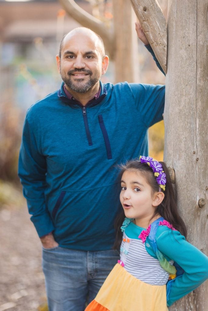 In Alexandria, Virginia, a man in a blue shirt leans against a tree, smiling, while a young girl in a colorful dress stands beside him, looking at the camera with an expressive face—capturing the perfect portrait moment.