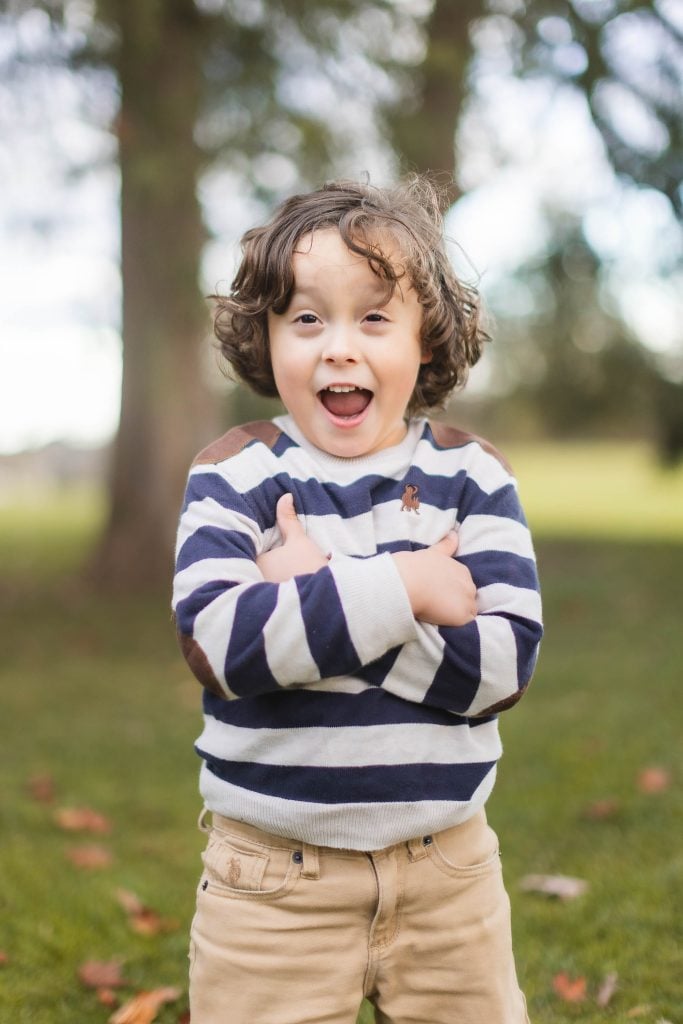 A young child with curly hair, wearing a striped sweater and beige pants, stands outdoors with arms crossed and an excited expression. Surrounded by trees and grass, it feels like a perfect family portrait moment captured at the Belair Mansion.