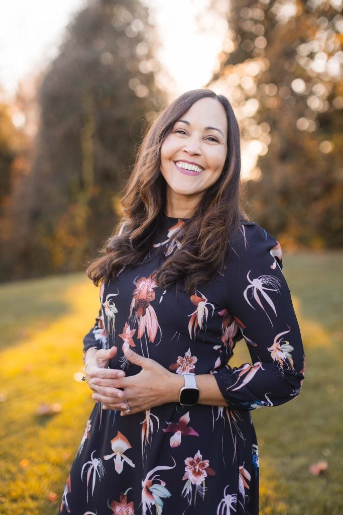 A woman with long brown hair, wearing a dark floral dress, stands outdoors on the grass at Belair Mansion. She smiles at the camera and touches her belly with both hands. The background includes trees and sunlight, creating a perfect scene for memorable portraits.