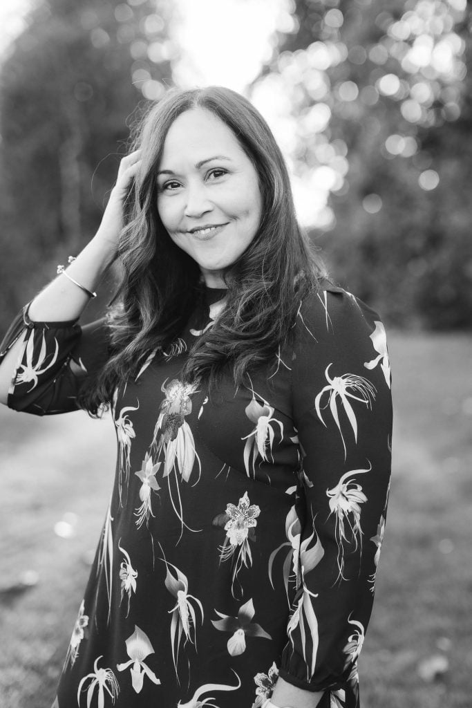 Woman with long hair wearing a floral dress smiles while touching her hair, standing outdoors with a blurred background of trees and grass. Black and white portrait taken at Belair Mansion.