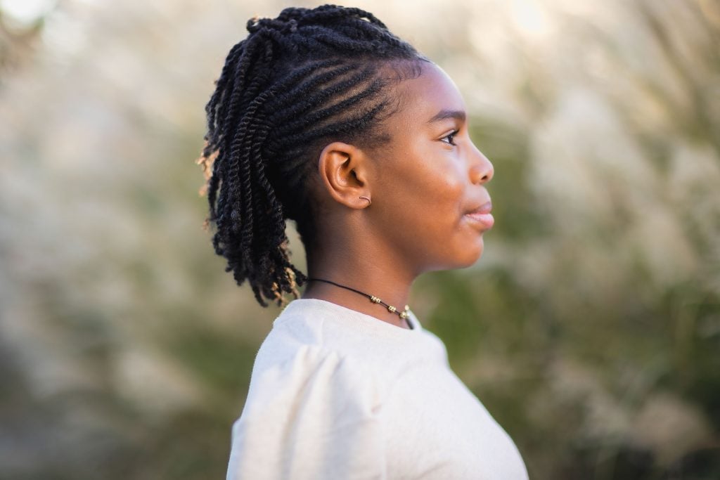 A person with braided hair stands outside, facing sideways. They are wearing a lightcolored top and a necklace, creating an intimate portrait that captures the essence of family moments.