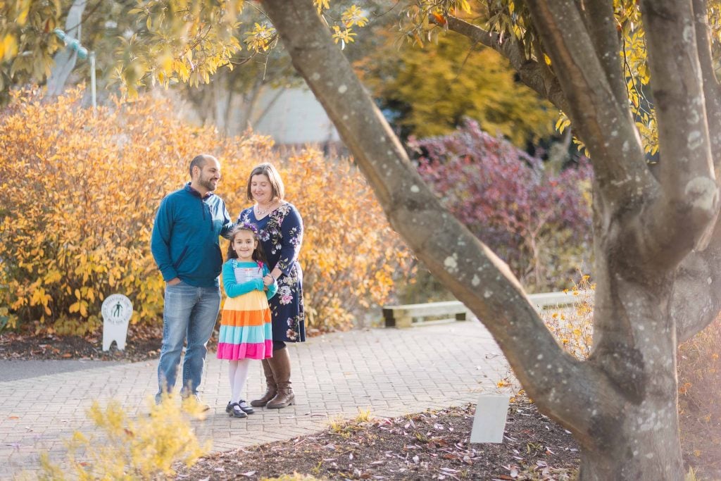 A man, woman, and young girl stand together on a paved path in Greenspring Garden during autumn, surrounded by colorful foliage and capturing a beautiful family portrait.