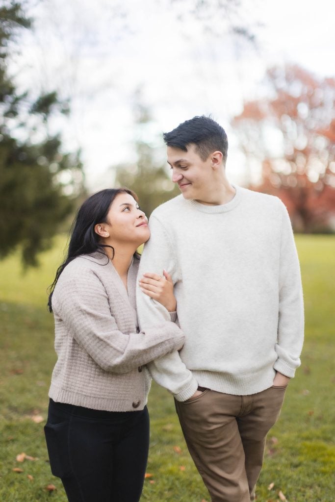 A couple stands outdoors near Belair Mansion, both wearing light sweaters. The woman holds the man's arm and looks up at him while he looks down at her, smiling. It's a perfect portrait moment, with trees and a lawn in the background.