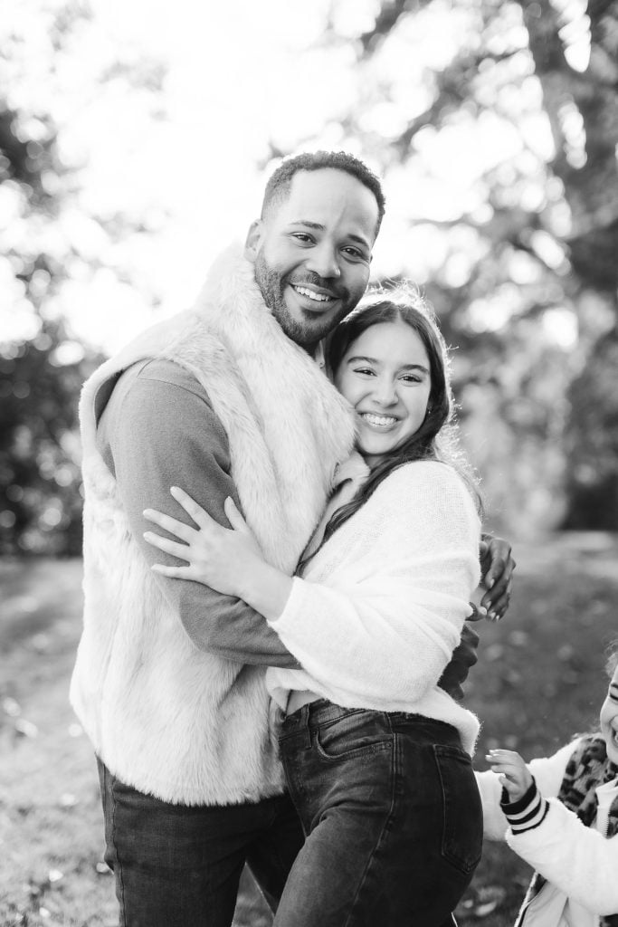 A man and a woman smile and hug outdoors, with trees in the background. The black and white portrait captures their joy, reminiscent of an extended family reunion at Belair Mansion.