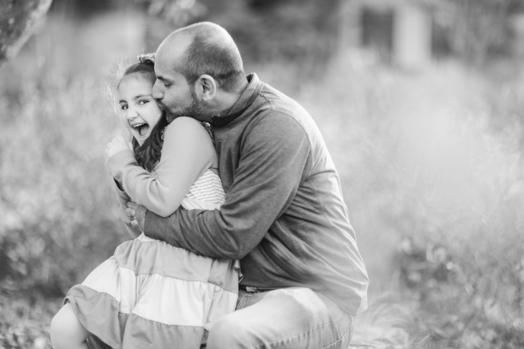 In this black and white portrait, a man hugs and playfully nibbles the ear of a smiling young girl. They are sitting outdoors in a blurred, natural setting, possibly in the serene surroundings of Alexandria, Virginia.