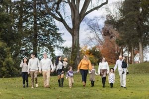 A family of ten, including adults and children, strolls across a grassy field with trees in the background, creating a picturesque portrait reminiscent of timeless moments at the Belair Mansion.