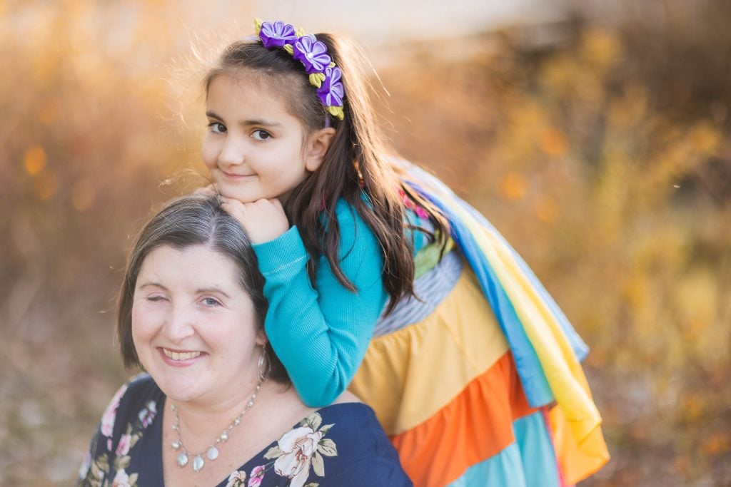 A woman with short hair and a floral top smiles while a young girl in a colorful dress and butterfly headband leans on her shoulder, capturing a lovely family moment at Greenspring Garden in Alexandria.